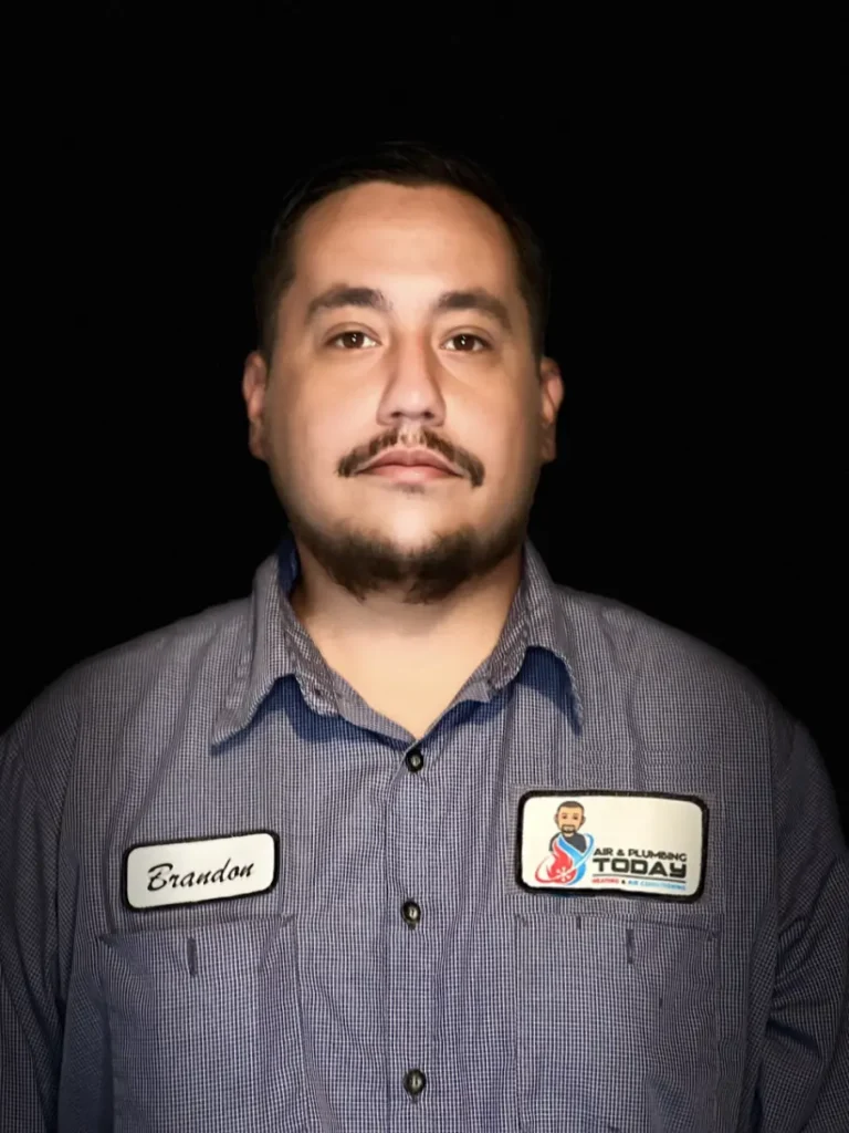 A man with short dark hair, a mustache, and a goatee wears a blue checkered work shirt with name tag "Brandon" and a company logo patch, standing against a black background for an about us profile photo.