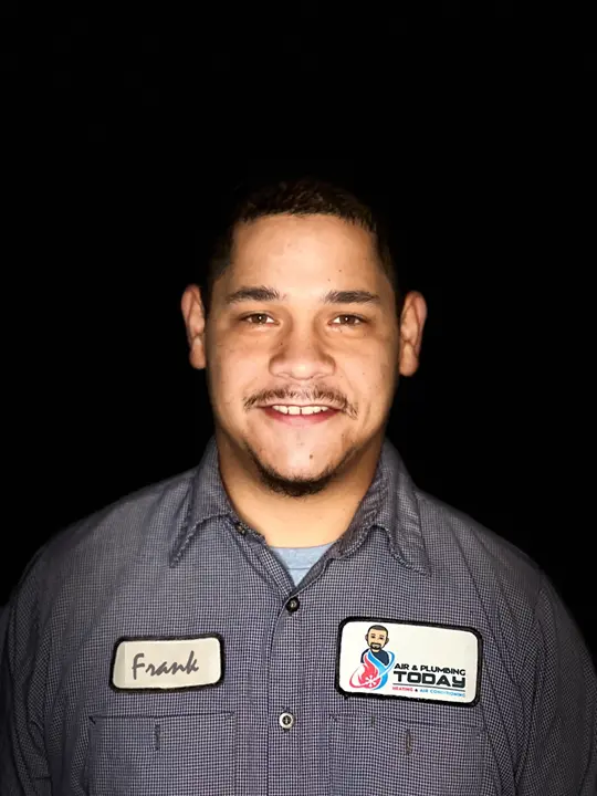 A man wearing a gray uniform shirt with the name tag "Frank" and an "A/C & Plumbing Today" logo stands against a black background, smiling at the camera—get to know our team on our about us page.