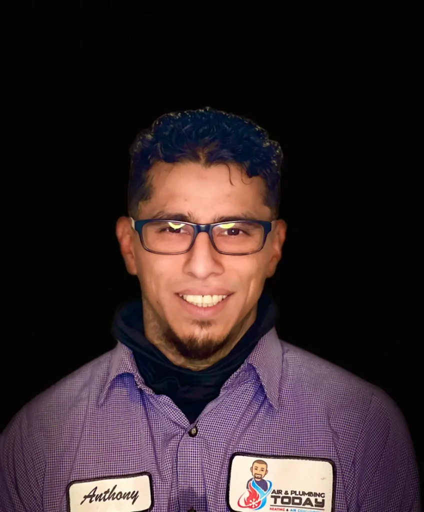 A man wearing glasses and a checkered shirt with a name tag "Anthony" and an "Air & Plumbing Today" patch smiles against a black background, reflecting the friendly team spirit highlighted in our about us section.