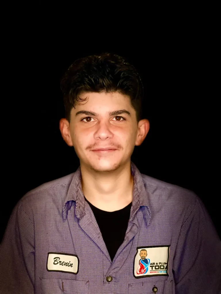 A young man with dark hair, wearing a checked work shirt and a name tag that reads "Brenin," poses against a plain black background, perfectly capturing the approachable spirit of our about us page.
