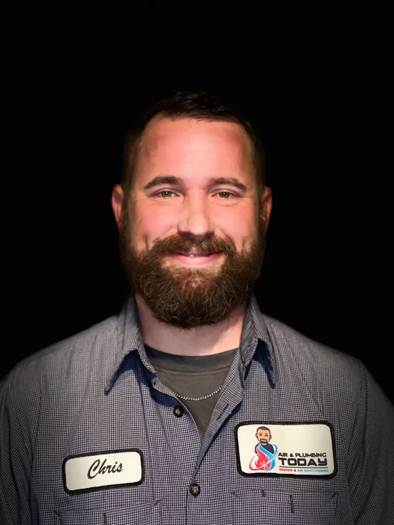 A man with a beard wearing a checkered shirt and "Chris" and "Air & Plumbing Today" name tags stands against a black background, smiling—ready to tell you more on our About Us page.