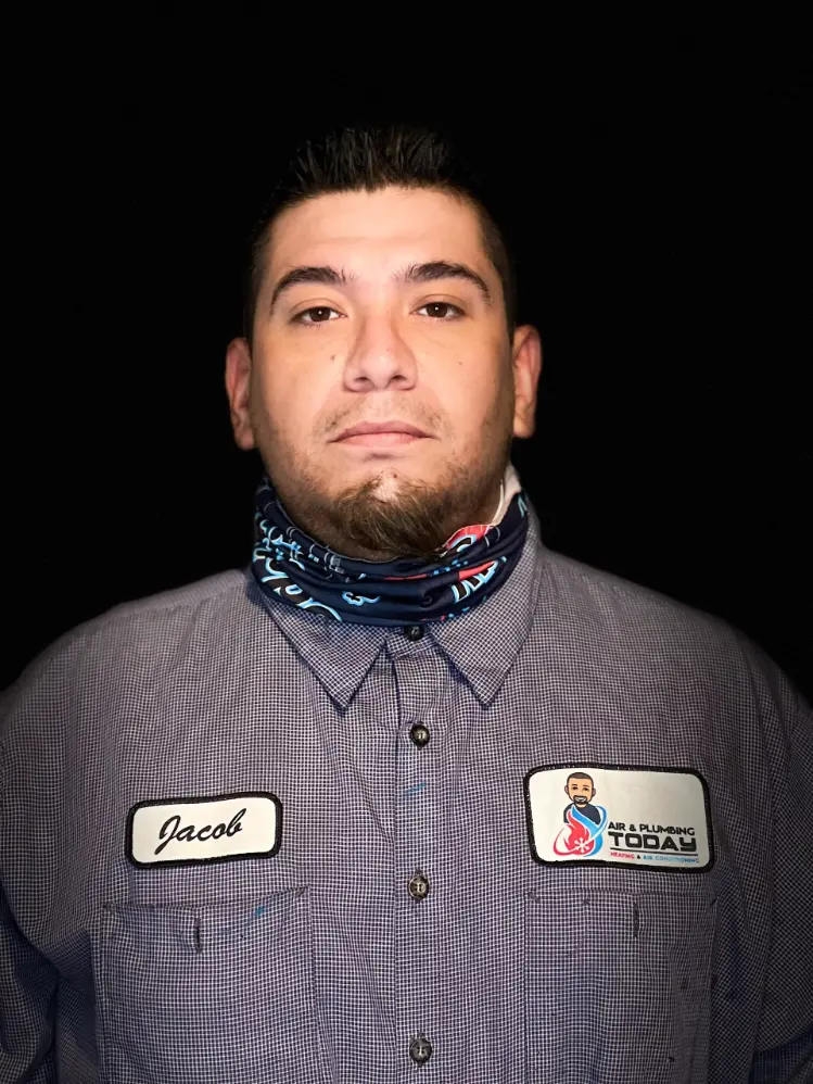 A man with short dark hair and a trimmed beard wears a checkered work shirt with a name patch "Jacob" and an "Air & Plumbing Today" logo, embodying our about us commitment, standing against a black background.