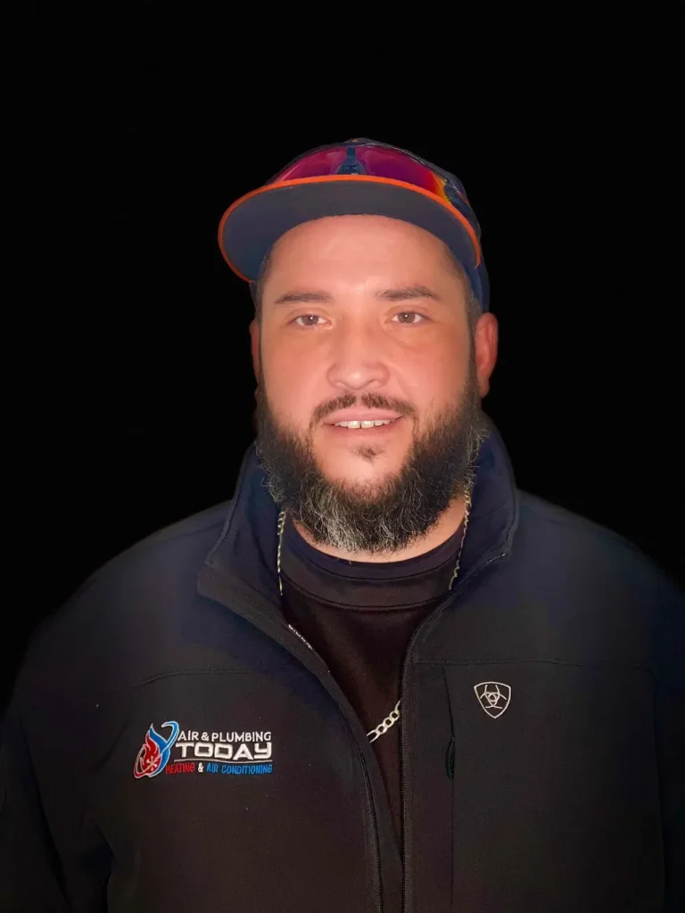A man with a beard and mustache wears a black jacket with a "Toby Air & Plumbing" logo and a black shirt, standing against a black background, representing the about us spirit of the company.