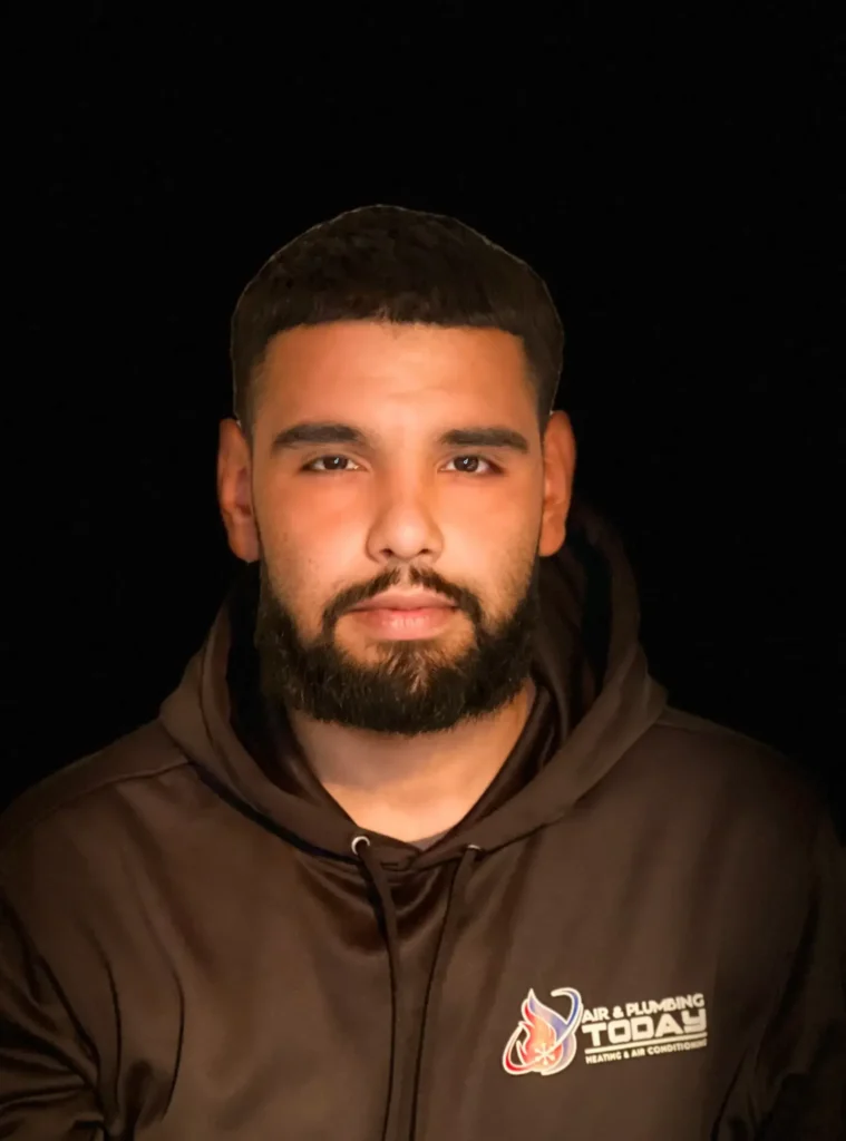 A man with a beard wearing a dark hoodie featuring the "Air & Plumbing Today" logo is posed against a black background, giving an authentic glimpse into our team for the about us section.