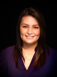 A woman with straight, dark hair and a purple top smiles at the camera against a plain black background, reflecting the approachable spirit of our About Us page.