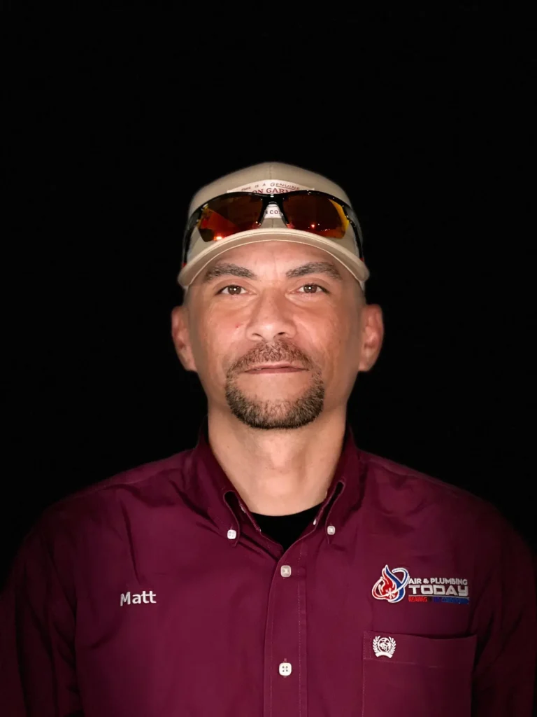 A man wearing a beige cap, sunglasses, and a maroon "Air & Plumbing Today" shirt with a name tag that reads "Matt," stands against a black background, representing our dedicated team in this about us snapshot.