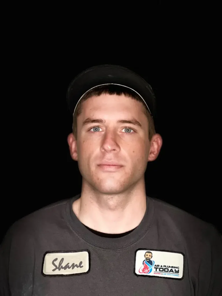 A man in a dark uniform and cap faces the camera against a black background. His shirt has a name tag reading "Shane" and an "Air & Plumbing Today" patch, reflecting the professionalism of our team featured on the about us page.