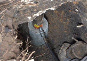 A hydraulic jack is installed under a house foundation during a foundation repair, surrounded by soil and exposed structural elements.