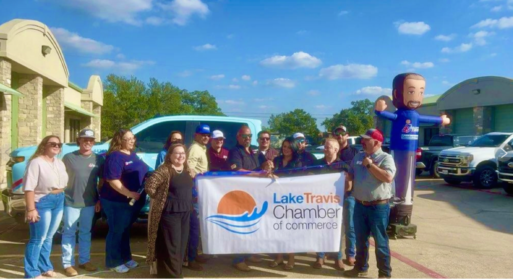 A group of people stand outdoors holding a "Lake Travis Chamber of Commerce" banner near trucks and a large inflatable figure under a sunny sky.