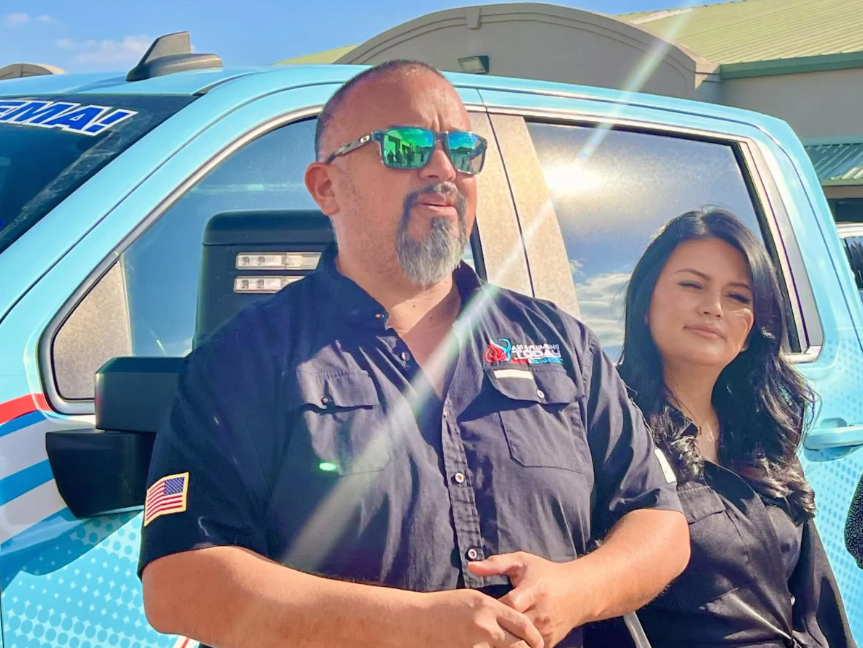 Two people stand in front of a blue vehicle on a sunny day. The man wears sunglasses and a shirt with an American flag patch; the woman stands beside him, both looking forward.
