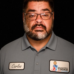 About Us: A man with glasses and a beard wears a gray polo shirt with name tag “Carlos” and an “Air & Plumbing Today” company logo, standing against a black background.