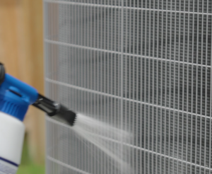 A person sprays water onto the coils of an outdoor air conditioning unit using a handheld spray bottle.
