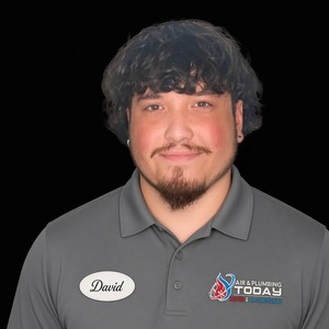 A man with short curly hair and a goatee wears a gray polo shirt with an “Air & Plumbing Today” logo and a name tag that says “David,” standing against a black background—an inviting image that perfectly represents the friendly team featured on our About Us page.