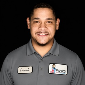 A man in a gray collared shirt with name tag “Frank” and a company logo stands against a black background, smiling at the camera—perfect for an “about us” introduction.