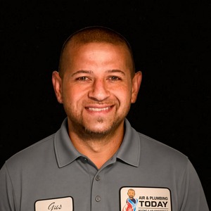 Man wearing a gray collared shirt with name tags stands against a black background, smiling at the camera, embodying the welcoming spirit of our About Us team.