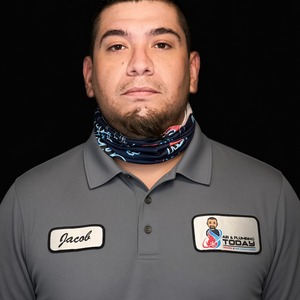 A man wearing a gray collared shirt with a name tag “Jacob” and a logo patch stands against a black background, wearing a patterned neck gaiter—capturing the hardworking spirit featured in our about us story.