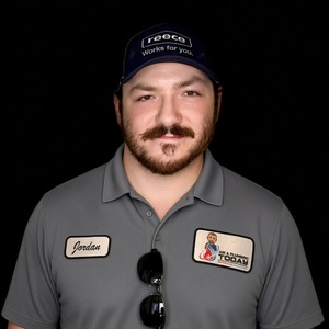 Man with a mustache wearing a dark cap and gray collared shirt with name tags, standing against a black background, representing our dedicated team featured on the About Us page.