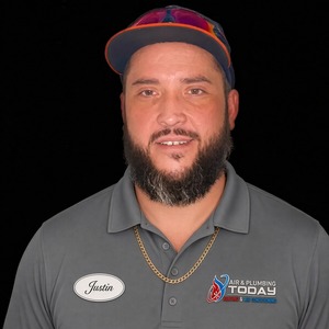 A man with a beard wearing a gray Air & Plumbing Today polo and a cap stands against a black background. His name tag reads “Justin,” representing the friendly professionals featured in our About Us section.