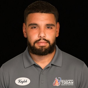 A man with short hair and a beard wears a gray “Air & Plumbing Today” polo shirt with a name tag reading “Kaylob” on a plain black background, representing the dedicated team featured on our About Us page.