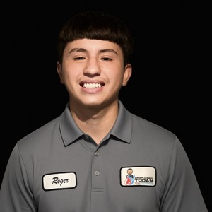 Young man with straight brown hair, wearing a gray collared shirt with a name tag that reads “Roger” and a company logo, smiling against a plain black background—perfect for an About Us page.