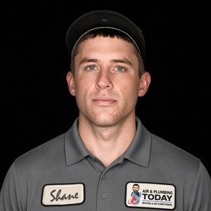 A man in a gray work shirt and cap stands against a black background. His shirt has badges with the name “Shane” and an “Air & Plumbing Today” logo, representing the dedicated team showcased on our About Us page.